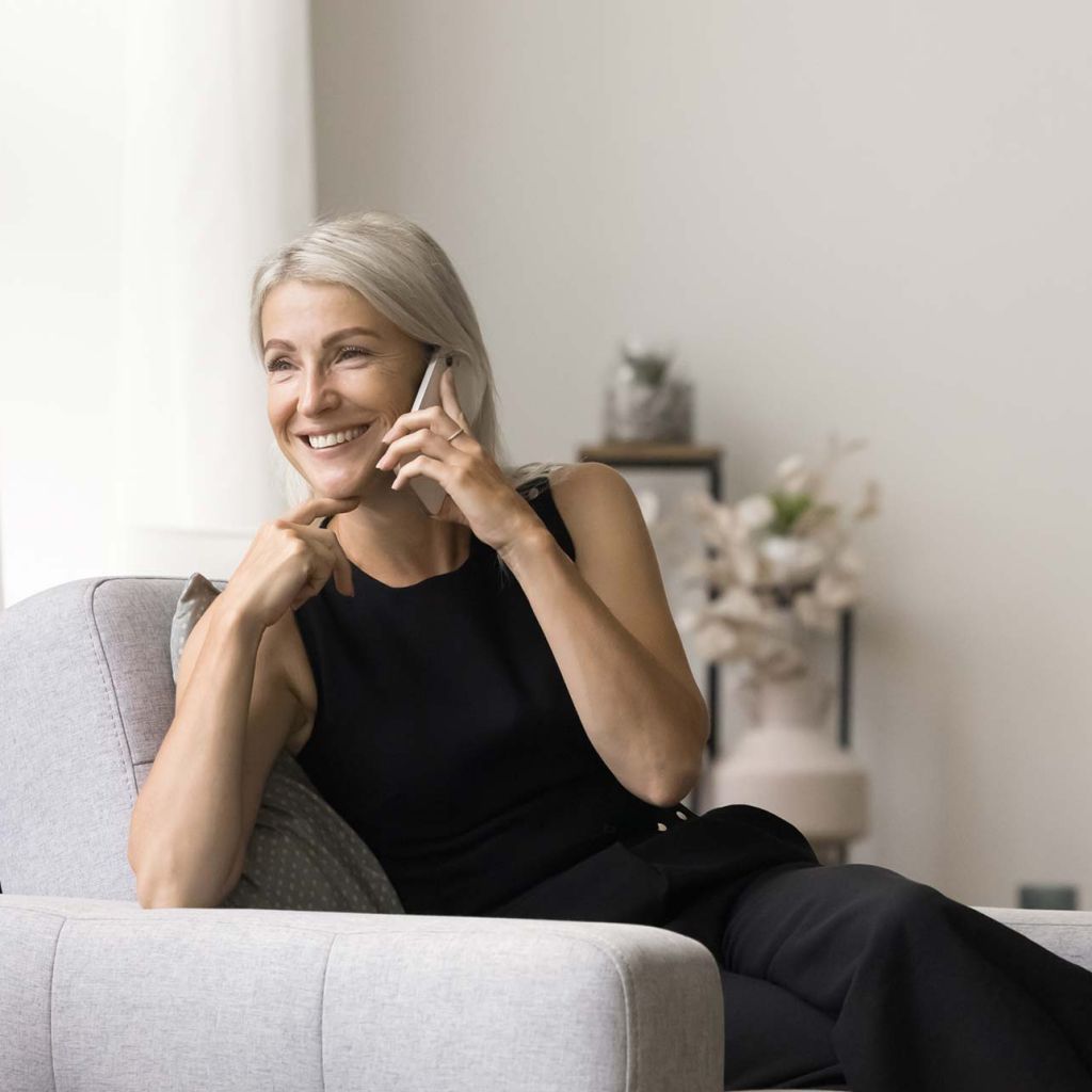 Woman talking on phone sitting on plush light gray chair in airy apartment